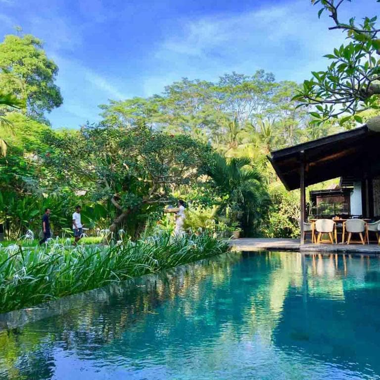 Surya Kembar Villas infinity pool with open-sided pavilion, dining seating, lush tropical vegetation and trees in background under blue sky