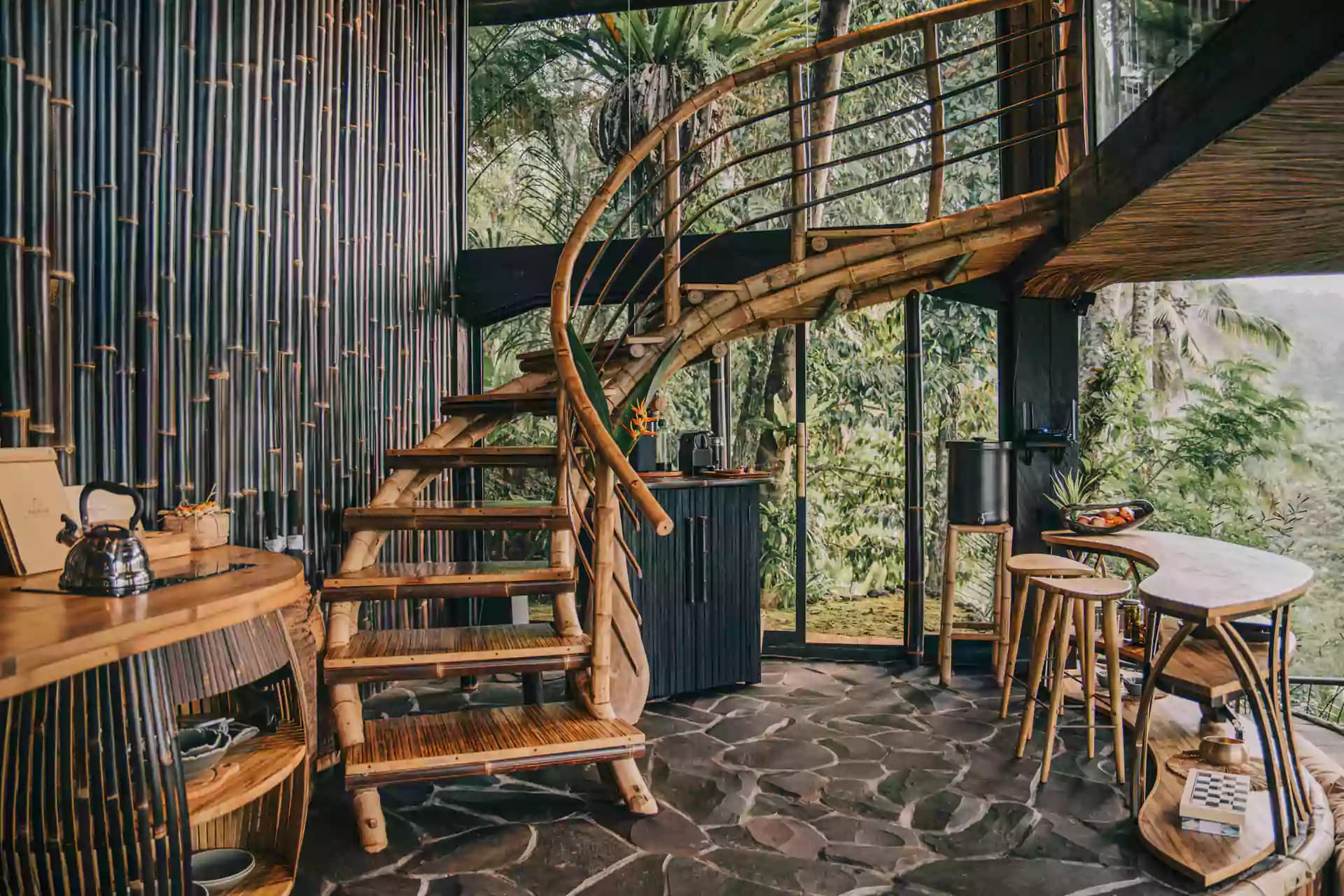 Interior of Hideout Eco Bamboo House Bali with wooden spiral staircase, dining table, desk, corrugated metal walls, and vegetation visible through windows