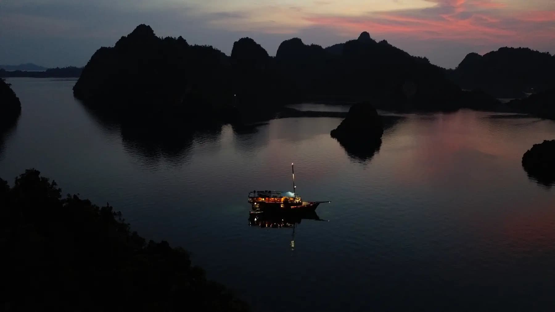 Sequoia liveaboard illuminated at dusk on calm water surrounded by silhouetted limestone karst formations under a pink sky