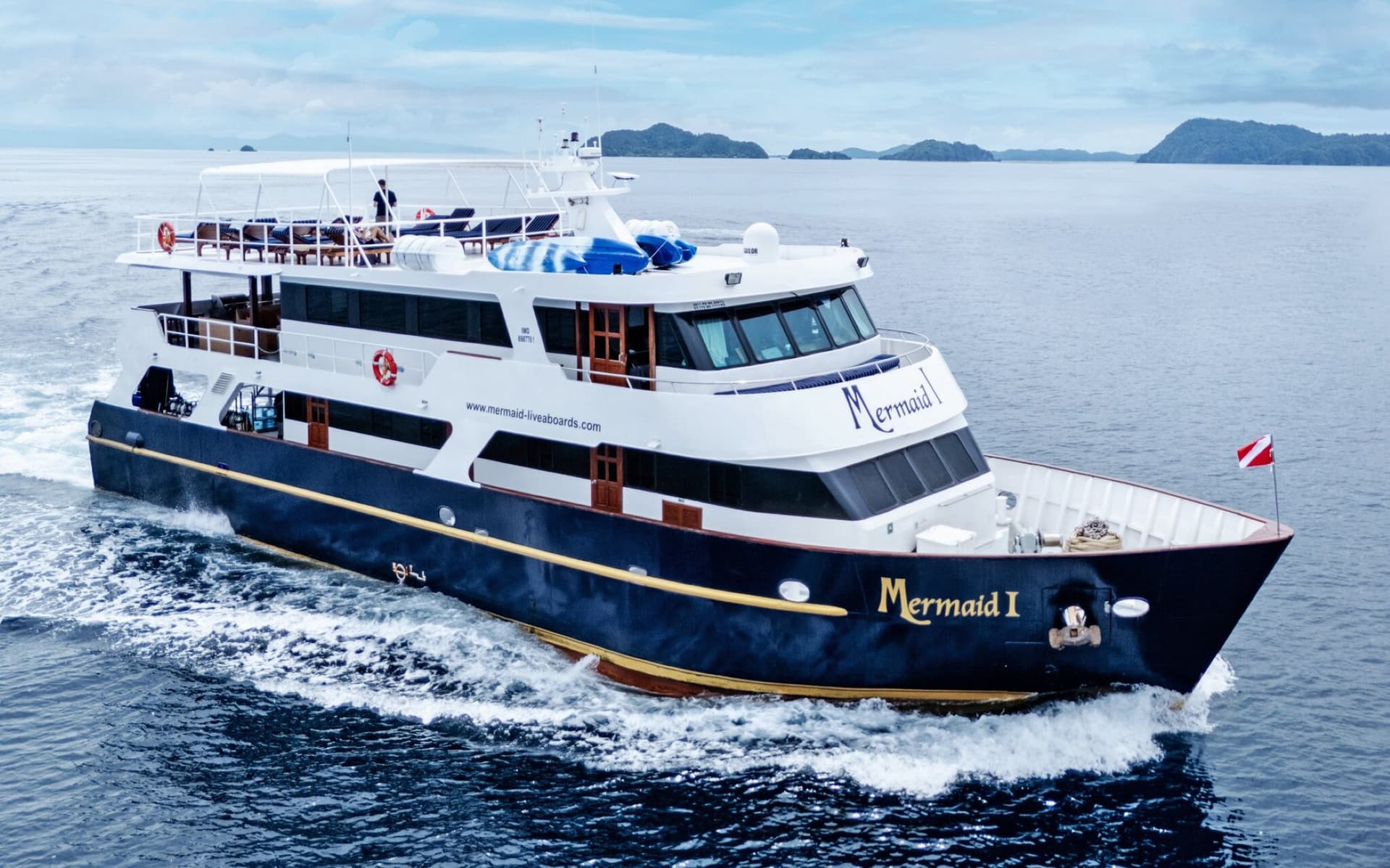 Mermaid I liveaboard cruising, dark blue hull and white superstructure, with a hilly coastline behind